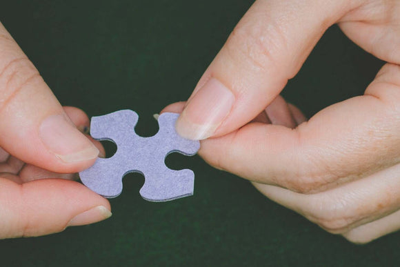 Close-up of a person's hands holding a purple jigsaw puzzle piece. The piece is shaped uniquely, showcasing intricate contours that fit together with other pieces. The background is dark, providing contrast to the hands and the puzzle piece, emphasizing the activity of assembling a jigsaw puzzle.