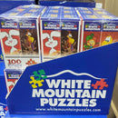A vibrant display of six mini cereal box puzzles from White Mountain Puzzles, featuring colorful cartoon characters on each box. Each puzzle contains 100 pieces, ideal for puzzle lovers and families. The boxes are neatly arranged in a blue packaging with the White Mountain Puzzles logo prominently displayed.