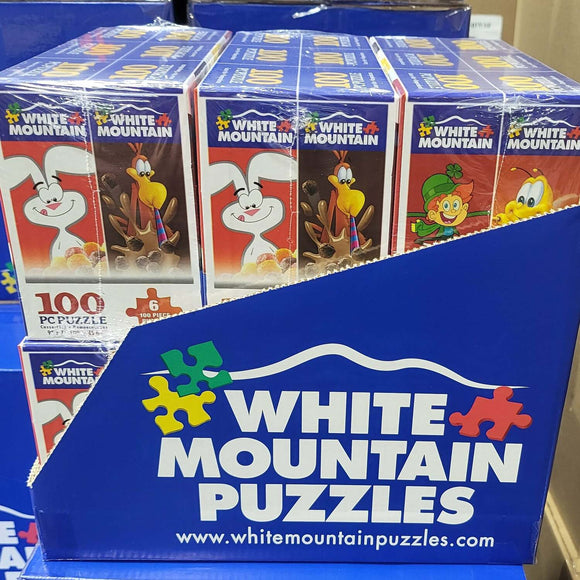 A vibrant display of six mini cereal box puzzles from White Mountain Puzzles, featuring colorful cartoon characters on each box. Each puzzle contains 100 pieces, ideal for puzzle lovers and families. The boxes are neatly arranged in a blue packaging with the White Mountain Puzzles logo prominently displayed.