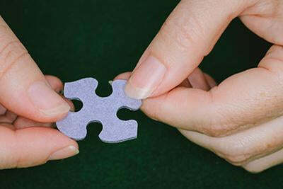 Close-up of hands holding a single jigsaw puzzle piece. The piece is gray with a unique four-way connector design. The background is green felt, adding a classic feel to the jigsaw puzzle experience.