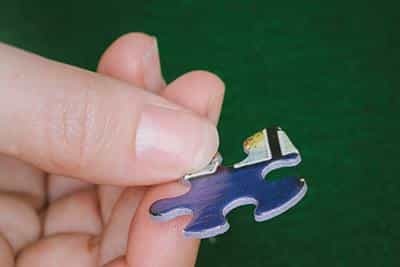 A close-up image of a hand holding a uniquely shaped puzzle piece from the 'Ice Cream Parlor' jigsaw puzzle by White Mountain Puzzles. The puzzle piece features vibrant colors and intricate details, indicative of an inviting ice cream theme.