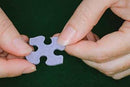 A close-up view of two hands holding a single jigsaw puzzle piece. The piece is purple and features a unique connector shape. The background is dark green, highlighting the textured surface of the puzzle piece, showcasing the tactile experience of assembling a puzzle.
