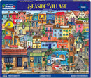 A colorful jigsaw puzzle box titled 'Seaside Village' by White Mountain Puzzles. The puzzle features a vibrant seaside town scene with various buildings, including a bakery, a patisserie, hotels, and a restaurant, all depicted in bright colors. The design includes people enjoying outdoor dining and lush green plants, capturing a cheerful, idyllic atmosphere. The box states it contains 500 larger pieces and includes a bonus poster inside.