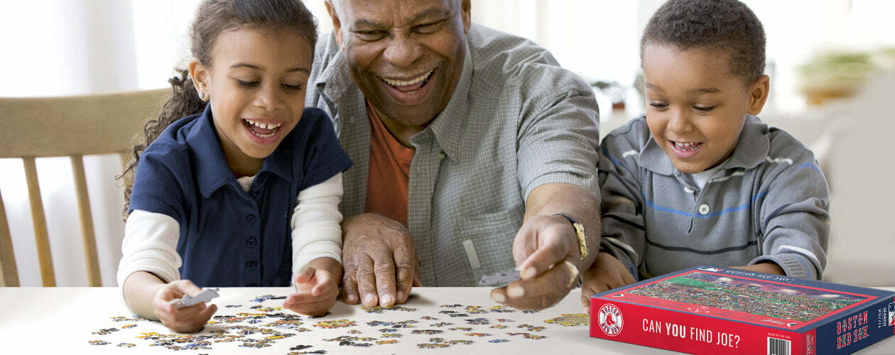 A cheerful scene featuring a grandfather and two children engaged in assembling a Boston Red Sox jigsaw puzzle by White Mountain Puzzles. The table is scattered with puzzle pieces, and the vibrant puzzle box is visible, showcasing fun and family bonding over a classic pastime.