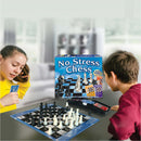 A girl and a boy sitting at a table playing No Stress Chess by Winning Moves. The game box is visible, featuring a vivid design. The chessboard is set up with black and white pieces, and the boy appears focused on the game, while the girl is holding a card and smiling. The background shows a cozy living room setting.