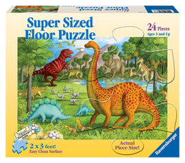 A colorful jigsaw puzzle box featuring a prehistoric landscape with various dinosaurs including a tall, spotted dinosaur, alongside a red T-rex and other species. The puzzle is designed for ages 3 and up and measures 2 x 3 feet. It contains 24 large pieces for easy handling, labeled with 'Actual Piece Size!' and 'Easy Clean Surface.'
