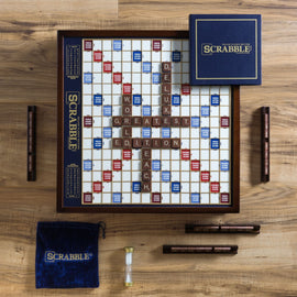Scrabble Deluxe Classic Edition game by Winning Solutions displayed on a wooden surface. The game board features traditional scoring with a mix of red and blue tiles indicating double and triple word scores. Labeled tiles spell out 'SCRABBLE', 'DELUXE', 'GREATEST', 'EDITION', and 'TEACH.' The game includes dark wooden letter tiles, a blue velvet tile pouch, an hourglass timer, and two score markers on the side.
