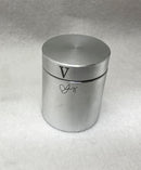 An aluminum cylinder featuring a smooth, polished surface with a simple, modern design. The top is flat with a small, circular indentation, and there is a decorative letter 'V' and an elegant signature on the side. The cylinder aims to intrigue and puzzle users with its engaging mechanical operation.