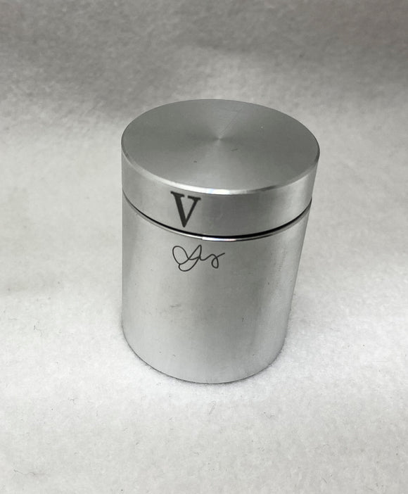 An aluminum cylinder featuring a smooth, polished surface with a simple, modern design. The top is flat with a small, circular indentation, and there is a decorative letter 'V' and an elegant signature on the side. The cylinder aims to intrigue and puzzle users with its engaging mechanical operation.