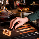 A hand moves a game piece on a beautifully crafted 15-inch Wenge replica backgammon set by Manopoulos. The game features rich wood tones and intricate design elements, with two dice nearby, and a glass of red wine in the background.