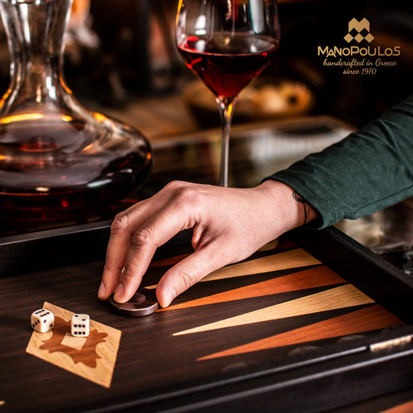 A hand moves a game piece on a beautifully crafted 15-inch Wenge replica backgammon set by Manopoulos. The game features rich wood tones and intricate design elements, with two dice nearby, and a glass of red wine in the background.