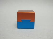 The Aluminum Dovetail Cube 02, designed by W.G.H. Strijbos, is a striking mechanical puzzle featuring a two-tone finish with a vibrant orange top and a contrasting blue base. Its smooth surfaces and unique dovetail design invite mechanical exploration, providing an engaging challenge for puzzle enthusiasts.