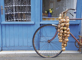 A vintage bicycle adorned with a cascade of golden onions, leaning against a blue wooden door with decorative curtains. The scene captures the charm of French markets and rustic decor.