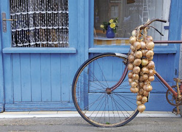 A vintage bicycle adorned with a cascade of golden onions, leaning against a blue wooden door with decorative curtains. The scene captures the charm of French markets and rustic decor.
