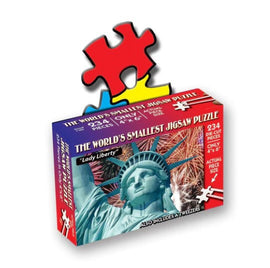 The world's smallest jigsaw puzzle featuring Lady Liberty with a vibrant packaging decorated in red, blue, and yellow. The box shows a photo of the Statue of Liberty surrounded by fireworks and includes a mention of tweezers for easy assembly. It contains 234 die-cut pieces that create a 4x6 inch puzzle when completed.