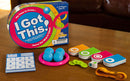 The game 'I Got This!' from Fat Brain Toys is displayed. The box features vibrant colors and the title prominently. Surrounding the box are various game pieces including three blue balls, colorful disc pieces, a pink ring, a scoring pad, and other game components, all set on a wooden table.