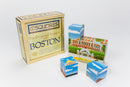 Boston City Squares jigsaw puzzle box featuring six original images of Boston landmarks. The puzzle pieces display vibrant illustrations of the State House and other iconic sites. The packaging is wooden with colorful graphics promoting Boston Folk Art.
