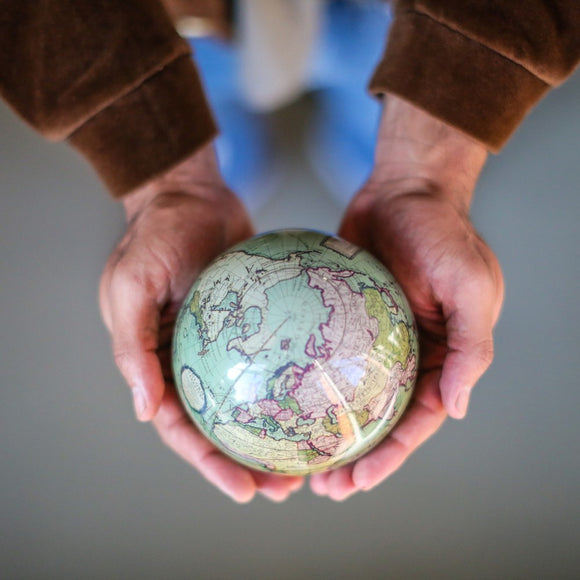 A person holding a 4.5-inch Mova Antique Terrestrial globe in their hands, showcasing detailed map designs with various colors and geographical features. The globe appears to have a smooth surface and reflects light, adding an elegant touch to its antique style.
