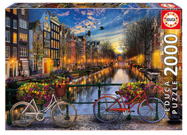 The AMSTERDAM WITH LOVE jigsaw puzzle by Educa features a scenic view of Amsterdam's iconic canals during sunset, with colorful bicycles adorned with flowers and charming traditional buildings lining the waterways, capturing the city's vibrant atmosphere in 2000 engaging pieces.