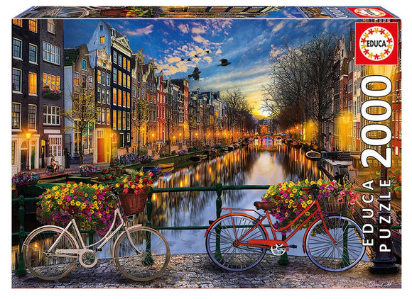 The AMSTERDAM WITH LOVE jigsaw puzzle by Educa features a scenic view of Amsterdam's iconic canals during sunset, with colorful bicycles adorned with flowers and charming traditional buildings lining the waterways, capturing the city's vibrant atmosphere in 2000 engaging pieces.