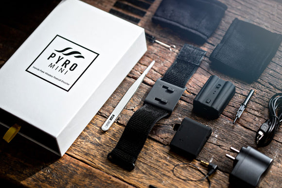 The Pyro Mini V4 Bundle by ELusionist laid out on a rustic wooden surface. The image features a white box with the Pyro Mini logo prominently displayed, along with various components including a wrist strap, battery pack, wires, an instruction tool, and two types of adapters.