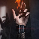 A hand with a tattooed wrist wearing a Pyro Mini V4 wrist device that emits flames. The device is black with a red light, set against a smoky backdrop. The user's fingers are poised as if performing a magic trick, showcasing the fiery effect created by the device.