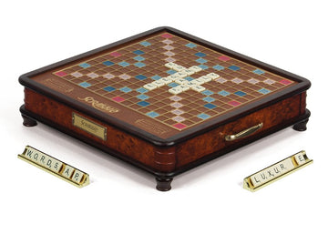 The Scrabble Luxury Edition game by Winning Solutions features an elegant wooden game board with a rich brown finish, displayed with a selection of letter tiles. The board shows various colorful squares for scoring, with some tiles already placed to form words like 'ELEGANT' and 'FAMILY'. There are also three tile racks on the side, holding letters that spell out 'WORDSAP' and 'LUXUR'.