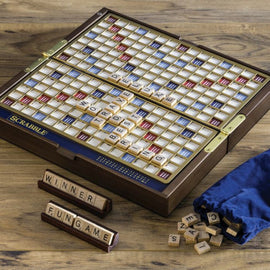 Scrabble Deluxe Travel Edition game displayed on a wooden table. The board is open with letters laid out, featuring a blue and gold design. Accompanying letter tiles are scattered nearby, along with a blue drawstring bag containing more tiles. A wooden tray holds the words 'WINNER' and 'FUNGAME'.
