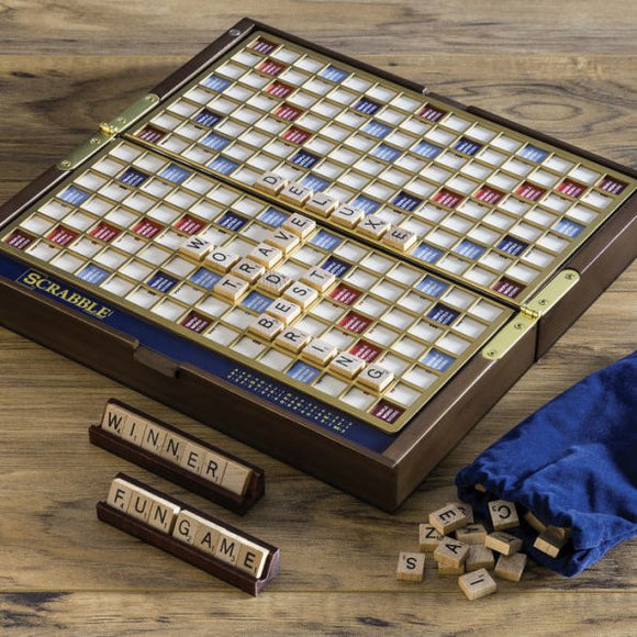 Scrabble Deluxe Travel Edition game displayed on a wooden table. The board is open with letters laid out, featuring a blue and gold design. Accompanying letter tiles are scattered nearby, along with a blue drawstring bag containing more tiles. A wooden tray holds the words 'WINNER' and 'FUNGAME'.