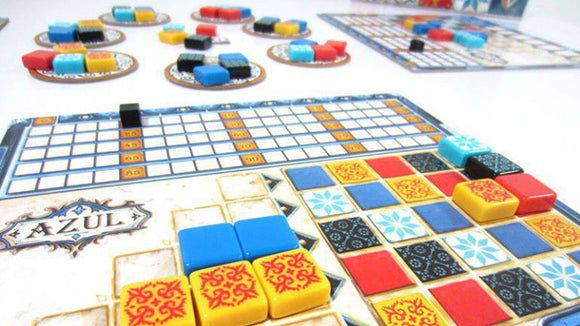 An image showcasing the game Azul by Plan B Games. The game features colorful tiles in red, blue, yellow, and black on player boards, with a central scoring area and multiple tile supply boards. Players can be seen arranging their tiles strategically in an engaging manner.