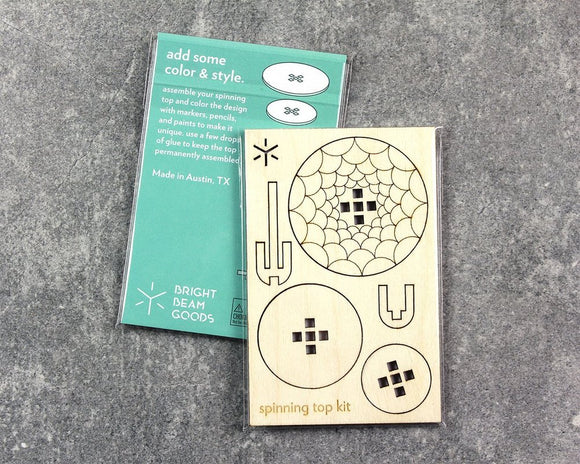 Spinning Tops Scales DIY kit by Bright Beam Goods, featuring a wooden board with various spinning top designs and a colorful instruction card, encouraging users to personalize their spinning tops with markers and paints. Perfect for creative hands-on fun.