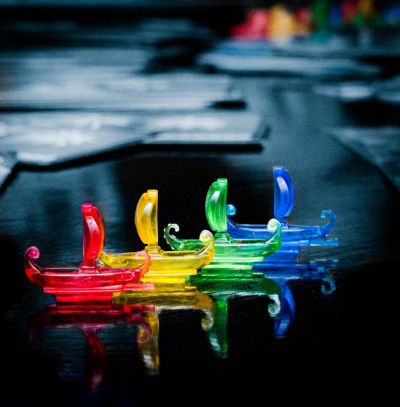 Colorful plastic ships in a row representing the game '7 Wonders: Armada' by Asmodee. Each ship is a different color: red, yellow, green, and blue, reflecting on a shiny surface. The game components are neatly arranged, showcasing the vibrant design and thematic elements of the game.