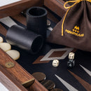 A close-up view of a 19-inch Backgammon game set by Manopoulous, featuring a fossil forest design. The game includes wooden pieces, two black dice, and a drawstring bag with the Manopoulous logo. The board showcases a stylish geometric pattern with smooth playing surfaces and brass accents, ideal for classic gameplay.