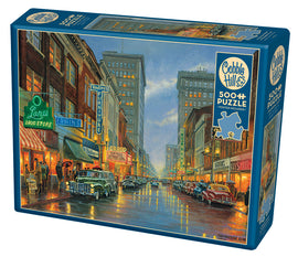 A Grand Night in Steubenville jigsaw puzzle by Cobble Hill featuring a nostalgic street scene illuminated by neon signs. The puzzle box showcases a vibrant, artistic illustration depicting a rainy night with vintage cars on the road, creating a warm, inviting atmosphere for puzzle enthusiasts.