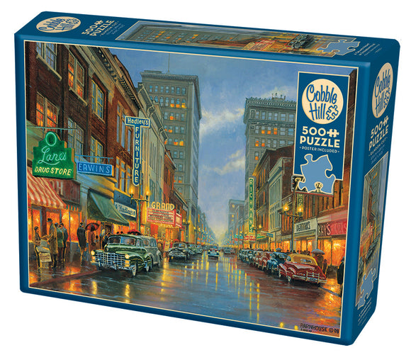 A Grand Night in Steubenville jigsaw puzzle by Cobble Hill featuring a nostalgic street scene illuminated by neon signs. The puzzle box showcases a vibrant, artistic illustration depicting a rainy night with vintage cars on the road, creating a warm, inviting atmosphere for puzzle enthusiasts.