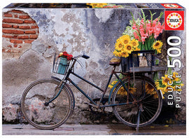 A beautifully designed 500-piece jigsaw puzzle featuring a vintage bicycle adorned with vibrant flowers. The scene captures the bike parked against a textured wall with a rustic look, showcasing a mix of colorful blooms that adds charm and life to the composition. Ideal for puzzle enthusiasts who appreciate floral art and nostalgic themes.