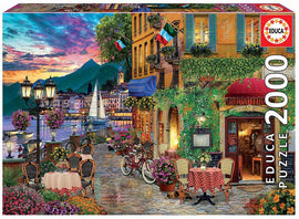 The ITALIAN FASCINO jigsaw puzzle by Educa features a vibrant street scene in Italy, showcasing colorful buildings, a beautiful sunset, and lively outdoor cafes adorned with flowers, bicycles, and traditional Italian decorations. This 2000-piece puzzle captures the charm of Italian culture and scenic landscapes.