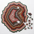 The Orbicular Geode Puzzle by Nervous System features a vibrant and intricately designed jigsaw puzzle. The puzzle depicts a geode with layers of colorful patterns in red, green, yellow, and brown, resembling a geological formation. Several puzzle pieces are scattered around the image, showcasing unique shapes that add to the overall aesthetic appeal of the puzzle. The outer edge of the puzzle is wavy and organic, enhancing its captivating design.