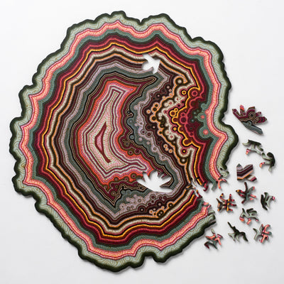 The Orbicular Geode Puzzle by Nervous System features a vibrant and intricately designed jigsaw puzzle. The puzzle depicts a geode with layers of colorful patterns in red, green, yellow, and brown, resembling a geological formation. Several puzzle pieces are scattered around the image, showcasing unique shapes that add to the overall aesthetic appeal of the puzzle. The outer edge of the puzzle is wavy and organic, enhancing its captivating design.