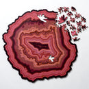 The Orbicular Geode Puzzle by Nervous System showcases a beautifully crafted jigsaw puzzle shaped like a geode slice. Featuring rich pinks and burgundy colors with intricate patterns, the puzzle is partially assembled, displaying several uniquely shaped pieces scattered around it. The design mimics the natural layers and textures found in geodes, providing a visually stunning and challenging experience for puzzle enthusiasts.