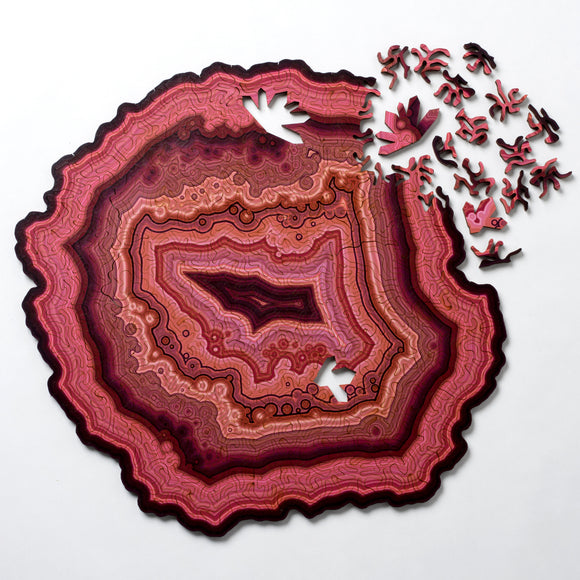 The Orbicular Geode Puzzle by Nervous System showcases a beautifully crafted jigsaw puzzle shaped like a geode slice. Featuring rich pinks and burgundy colors with intricate patterns, the puzzle is partially assembled, displaying several uniquely shaped pieces scattered around it. The design mimics the natural layers and textures found in geodes, providing a visually stunning and challenging experience for puzzle enthusiasts.