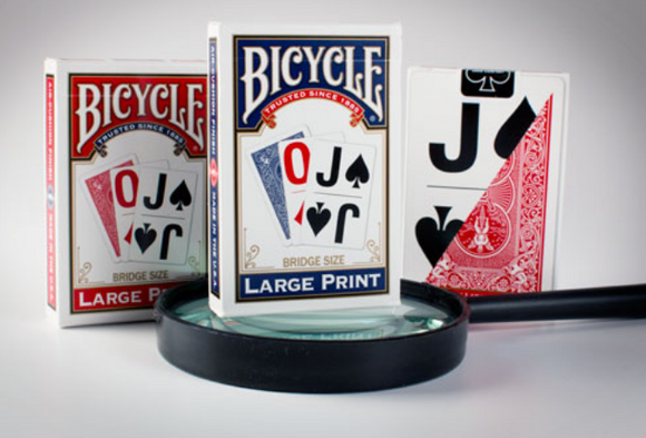 Bicycle Bridge Size Large Print playing cards displayed in a close-up shot, featuring a red and blue design. The cards are shown through a magnifying glass, highlighting the large print for easy reading. Two additional decks are partially visible in the background, emphasizing the vibrant colors and recognizable Bicycle branding.