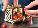 A wooden mechanical DIY kit called UGears 3D: Dice Keeper. The intricately designed box features gears inside and is shown with hands placing colorful dice into it. The background is a dark surface, enhancing the focus on the product.