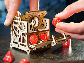 A wooden mechanical DIY kit called UGears 3D: Dice Keeper. The intricately designed box features gears inside and is shown with hands placing colorful dice into it. The background is a dark surface, enhancing the focus on the product.
