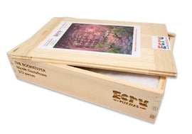 The Bookkeeper jigsaw puzzle by Nicole Gustafsson, produced by Artifact Wooden Puzzles. The image shows a beautifully crafted wooden box containing the puzzle with a colorful artwork of a whimsical setting on the cover. The packaging features the product's title and brand laser-etched onto the wood, showcasing its quality and craftsmanship.