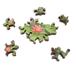 A detailed view of the jigsaw puzzle 'Cathedral of the Changing Tides' by Aaron Wolf. Several wooden puzzle pieces are scattered around, showcasing a vibrant design featuring green leaves and a delicate pink flower. The puzzle pieces are intricately shaped, reflecting high-quality craftsmanship by Artifact Wooden Puzzles.