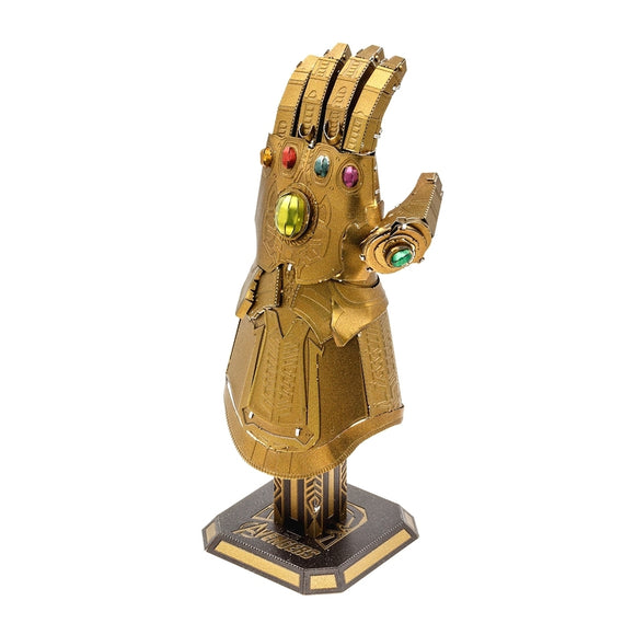 A detailed metal model of the Infinity Gauntlet from the Avengers, featuring intricate designs and colorful gem-like embellishments. The gauntlet is gold-colored and stands on a decorative base, showcasing the craftsmanship of the DIY kit by Fascinations.
