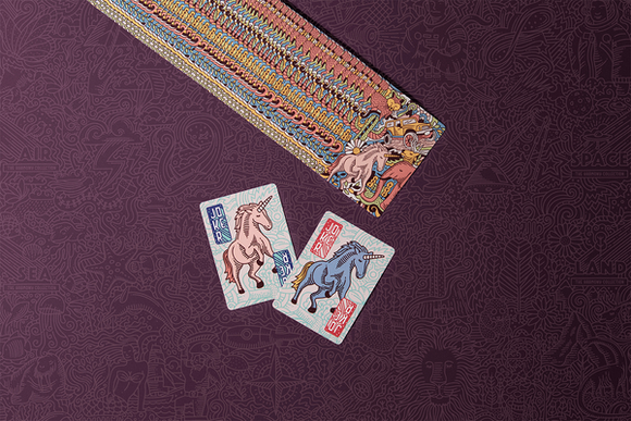 The Harmony Collection: Land by US Playing Card Co. features colorful cards with whimsical designs, including unicorns, set against a richly patterned purple background. The image shows a horizontal row of cards adorned with artistic illustrations, emphasizing creativity and fun.
