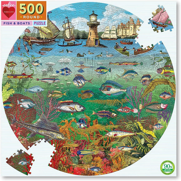 A colorful 500-piece round jigsaw puzzle titled 'Fish & Boats' by Eeboo. The image features a vibrant underwater scene with various fish, coral, and an idyllic coastal view showcasing sailing boats and a lighthouse. A piece of the puzzle is shown detached in the top right corner.