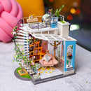 Dora's Loft DIY Model Kit by Robotime is a delightful miniature home model featuring intricate details. It showcases a spiral staircase, cozy living area with bookshelves, a soft pink chair, and vibrant plants. The scene is crafted with artistic touches and realistic furnishings, making it perfect for craft enthusiasts and model builders.